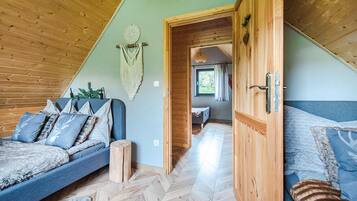 Een knus slaapkamer in loftstijl met een houten plafond, een groot bed met beddengoed met een patroon, een klein zithoekje en zicht door een deuropening naar een andere kamer.