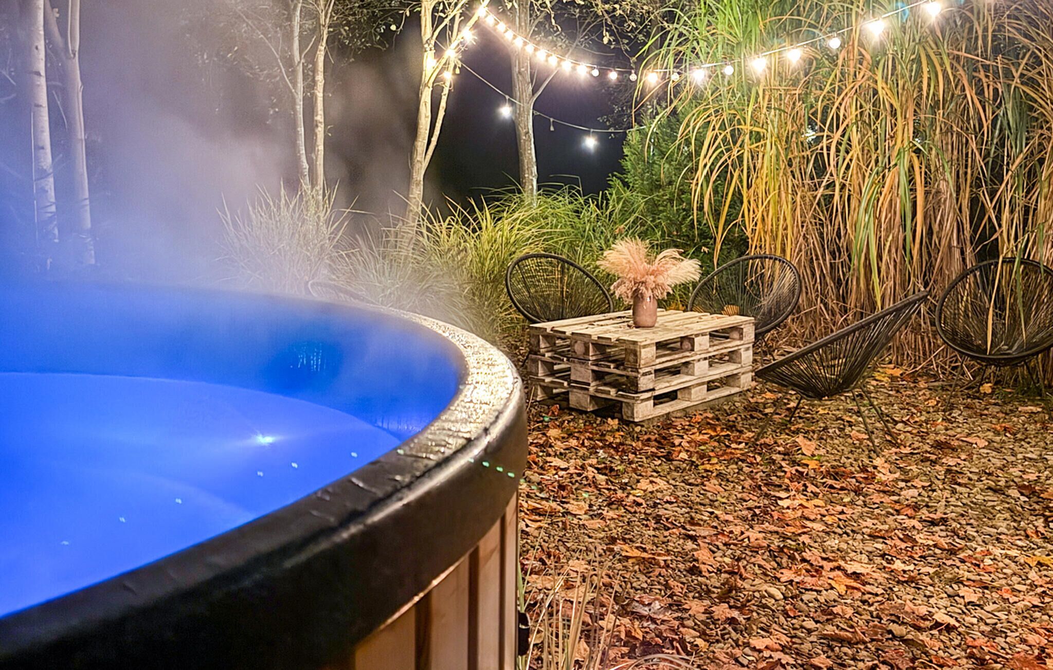 Een hottub omgeven door een rustieke buitentoevlucht met een houten tafel en stoelen, sfeerverlichting en hoge grassen.