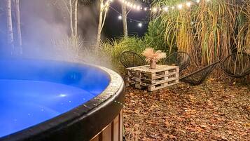 Een hottub omgeven door een rustieke buitentoevlucht met een houten tafel en stoelen, sfeerverlichting en hoge grassen.