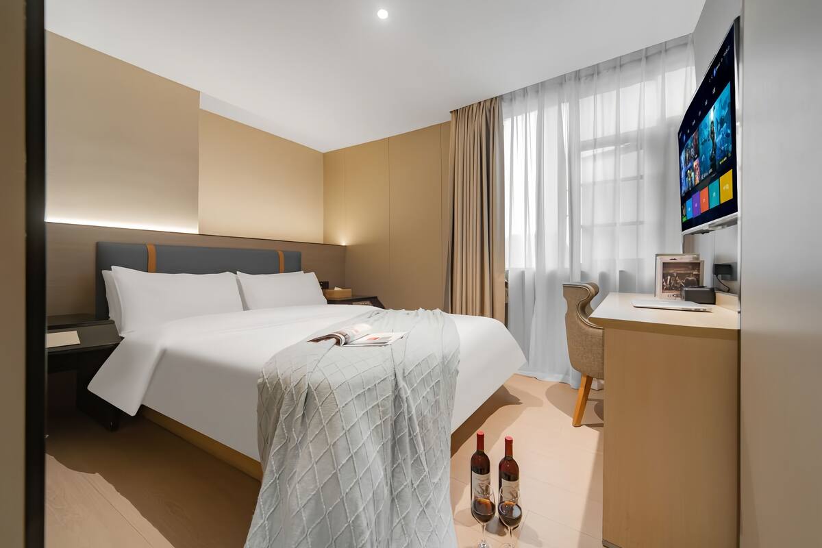 A hotel room with a bed, a desk, a TV, and two bottles of wine.