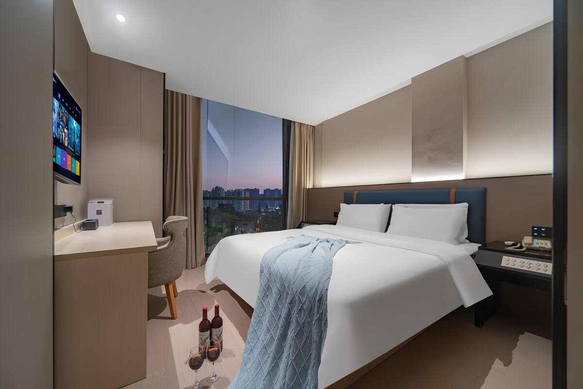 A modern hotel room with a large bed, a desk, a chair, a nightstand, and a TV mounted on the wall.