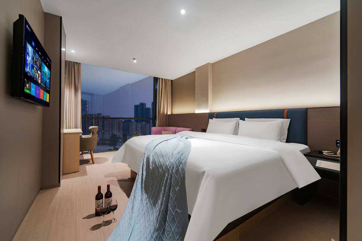 A modern hotel room with a large bed, a TV mounted on the wall, a desk with a chair, and a view of the cityscape.