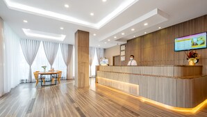Zona de recepción de un hotel con un mostrador de madera, un área de espera con una mesa y sillas, y una pantalla grande que muestra información.