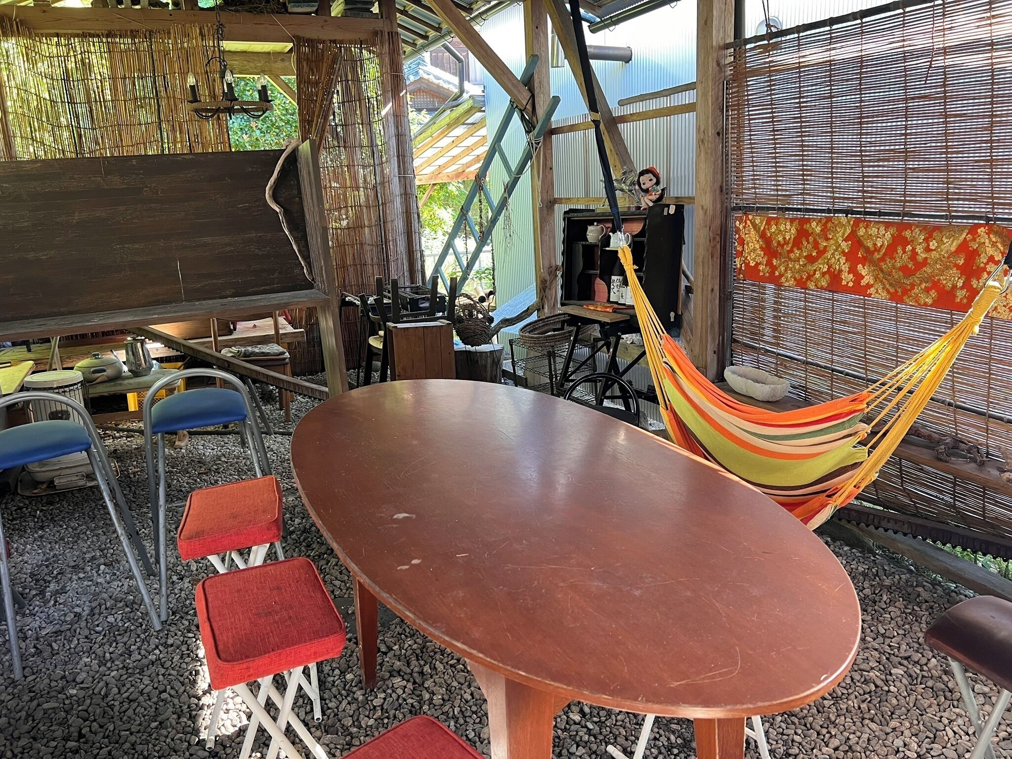 An outdoor seating area with a wooden table and red chairs, a hammock, and a bamboo wall.