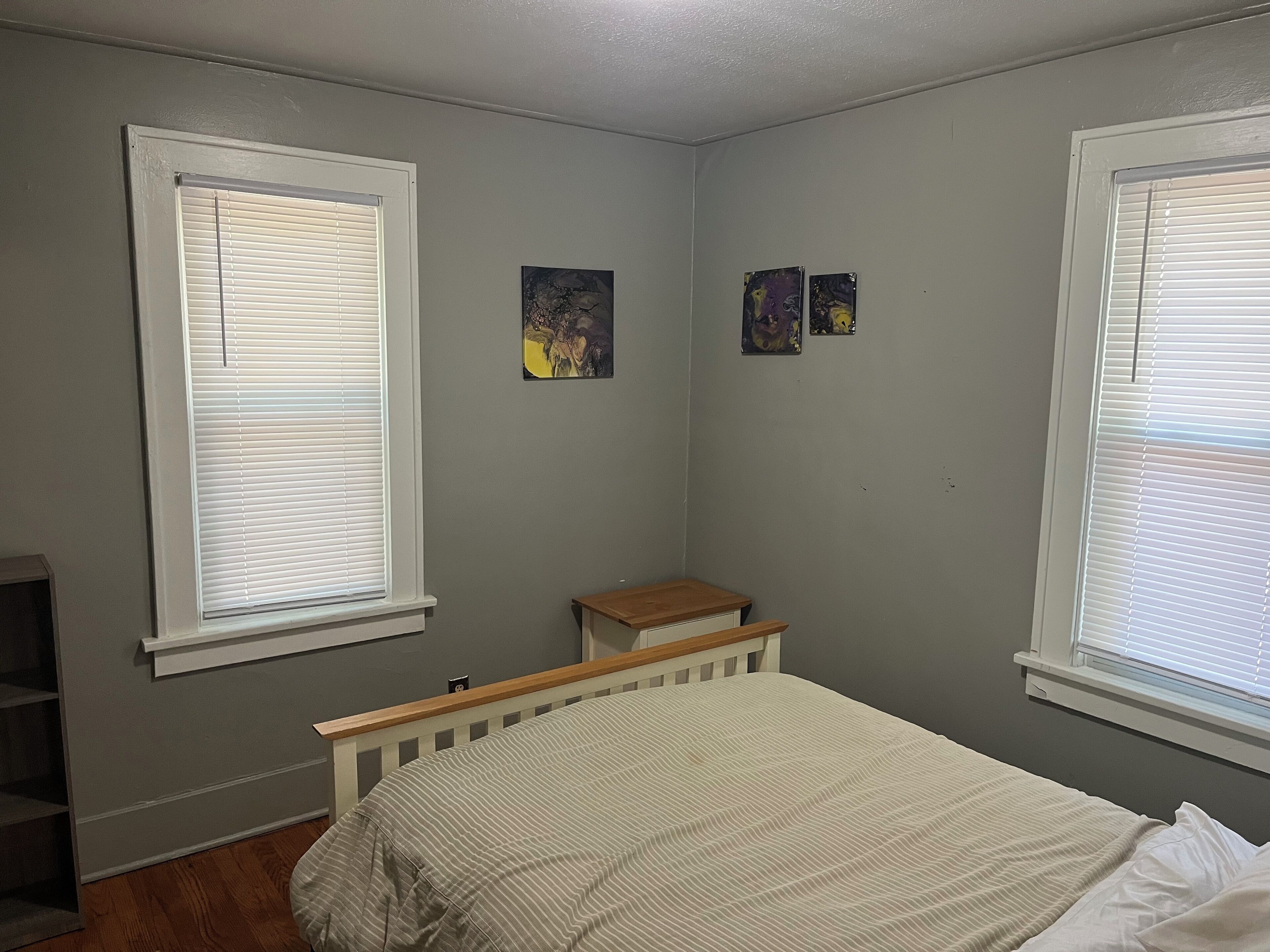 A bedroom with two windows, a bed, a nightstand, and two wall-mounted paintings.