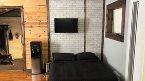 Makuuhuoneessa on sänky, tiiliseinään asennettu televisio, juomavesiautomaatti ja puinen penkki.