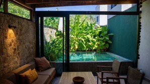 A cozy outdoor seating area with a stone wall, a wooden table, and a view of a swimming pool surrounded by lush greenery. - Casa Premium com Piscina e rea Gourmet - Camburi (São Sebastião)