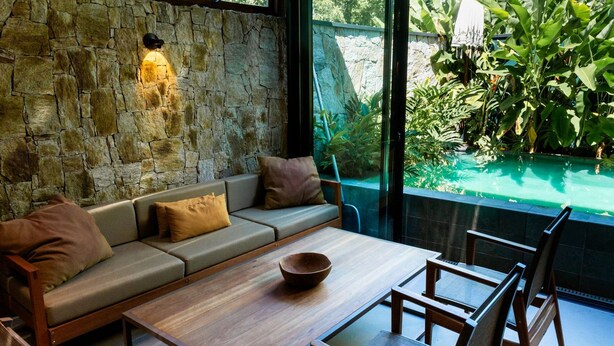 A modern interior with a stone wall, a sofa, a wooden table, and a view of a pool and greenery outside. - Casa Premium com Piscina e rea Gourmet - Camburi (São Sebastião)