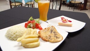 Une table à manger avec une assiette de nourriture comprenant du riz, des frites, une tranche de viande et un dessert, ainsi qu'un verre de jus d'orange.