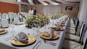 Une longue table de banquet, recouverte de nappes blanches, agrémentée d’assiettes dorées et de verres transparents.