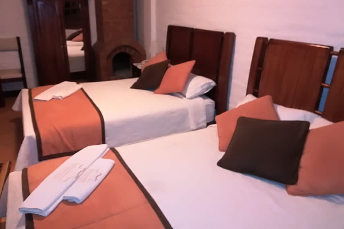 Ein Hotelzimmer mit zwei Einzelbetten, jeweils mit weißer Bettdecke und orangefarbenen Kissen. Im Hintergrund ist ein Holzschrank und ein Kamin aus Ziegelsteinen zu sehen.