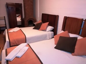 Habitación de hotel con dos camas individuales, cada una con una colcha blanca y almohadas naranjas. Se observa un armario de madera y una chimenea de ladrillo al fondo.