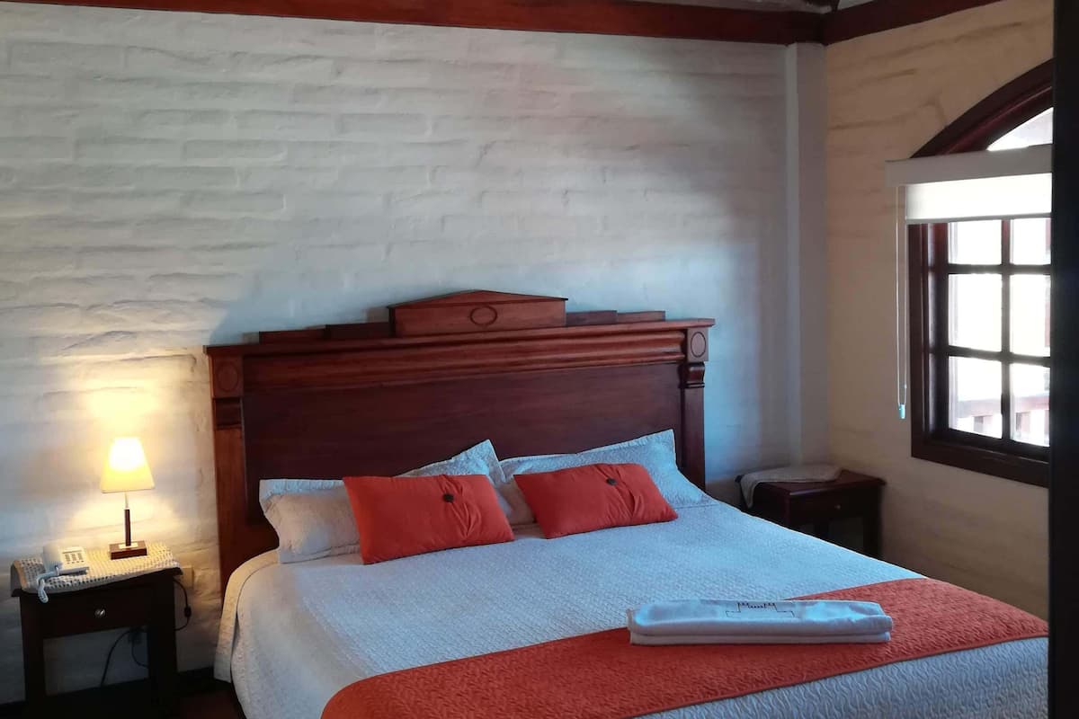 Ein Schlafzimmer mit Holzbalkendecke, einem Bett mit weißer und orangefarbener Bettwäsche, einem Nachttisch mit Lampe und einem Fenster mit Blick.