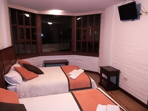 Habitación de hotel con dos camas, una mesita pequeña, un televisor montado en la pared y ventanas amplias con vistas al exterior.