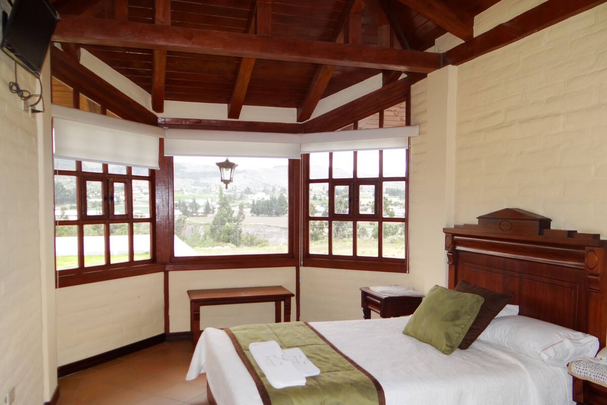 Ein Schlafzimmer mit hölzernem Kopfteil, einem Bett mit grün-weiß gestreifter Bettdecke, einem Nachttisch und einem Fenster mit Blick ins Freie.