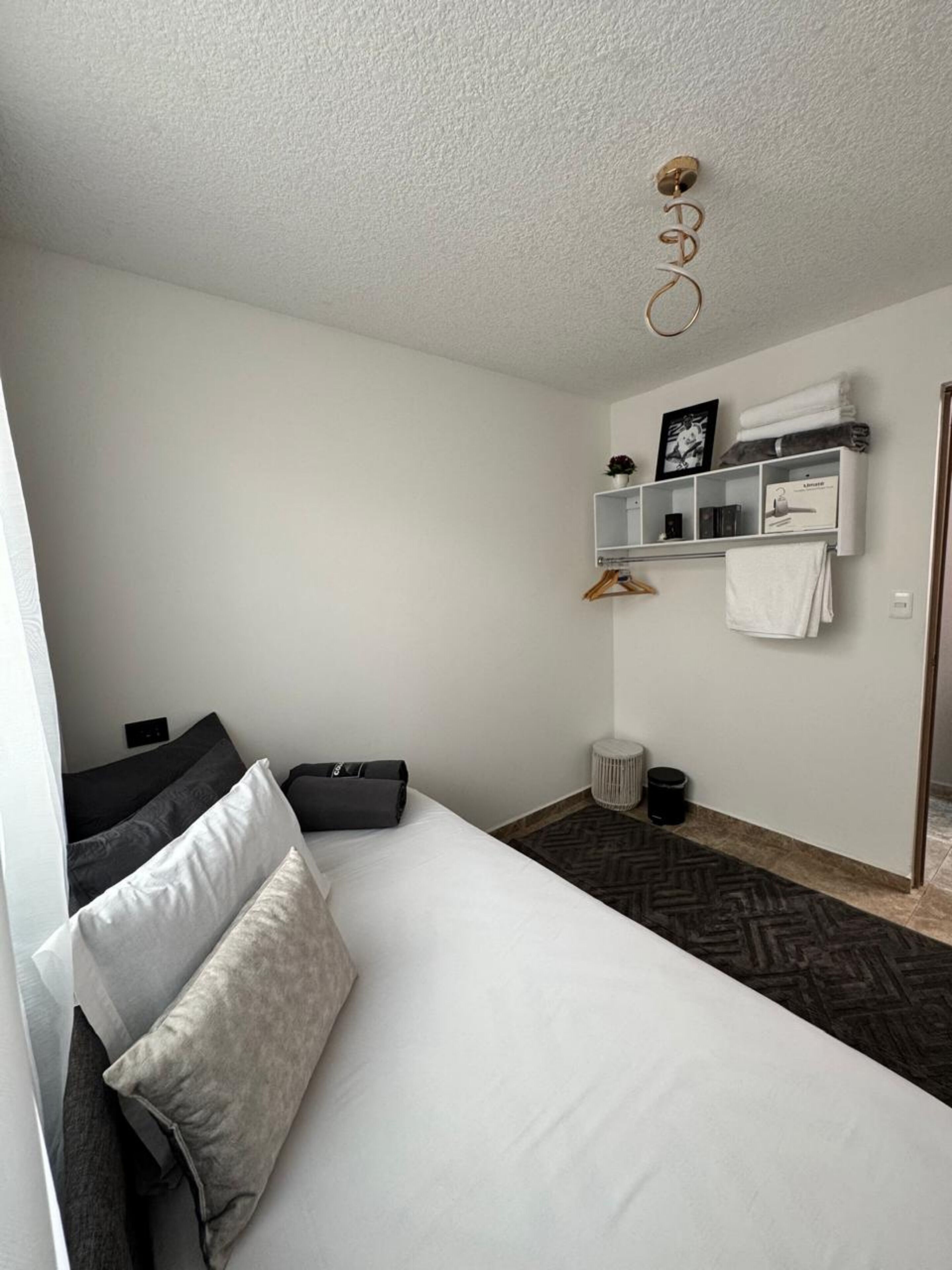 Habitación de hotel minimalista con una cama individual, una mesita de noche pequeña, un estante en la pared con toallas y una lámpara en el techo.