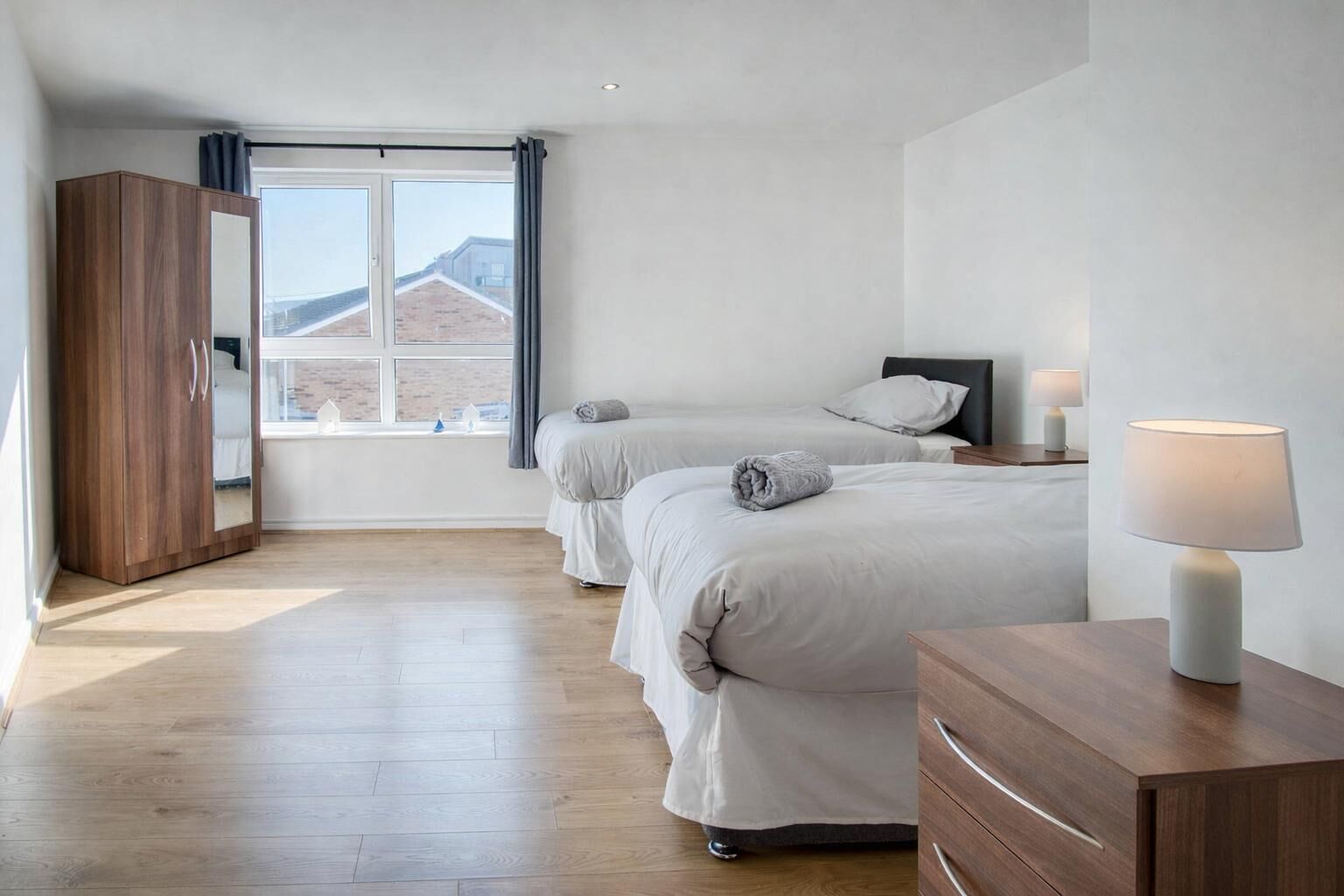 A bedroom with two beds, a wardrobe, a bedside table with a lamp, and a window with curtains.