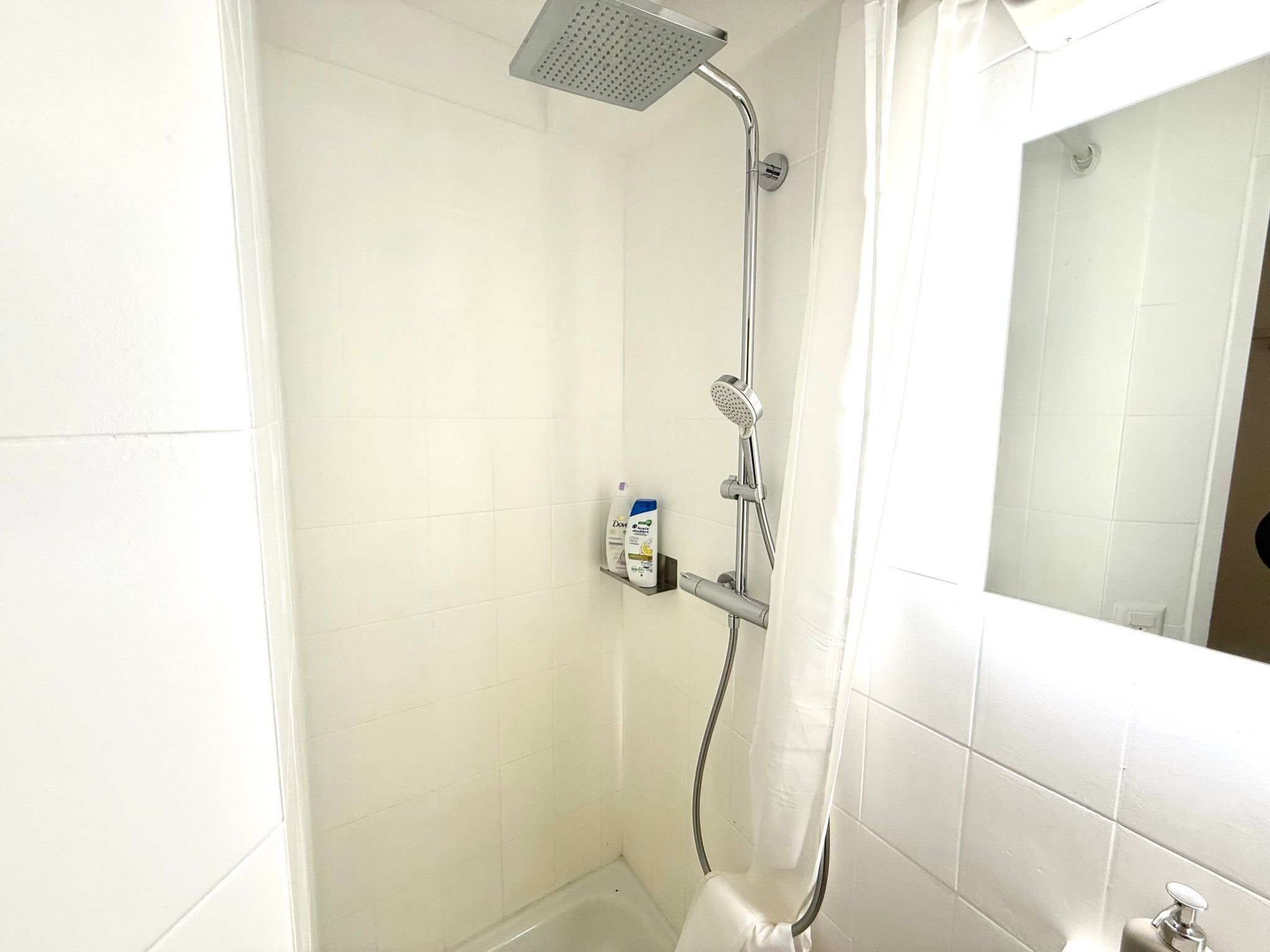 Une petite salle de bain aux murs recouverts de carreaux blancs, équipée d’une douche, de toilettes et d’un miroir.