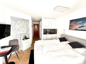 Pieni hotellihuone, jossa on sänky, työpöytä, tuoli, televisio, pieni keittiö ja kylpyhuone.