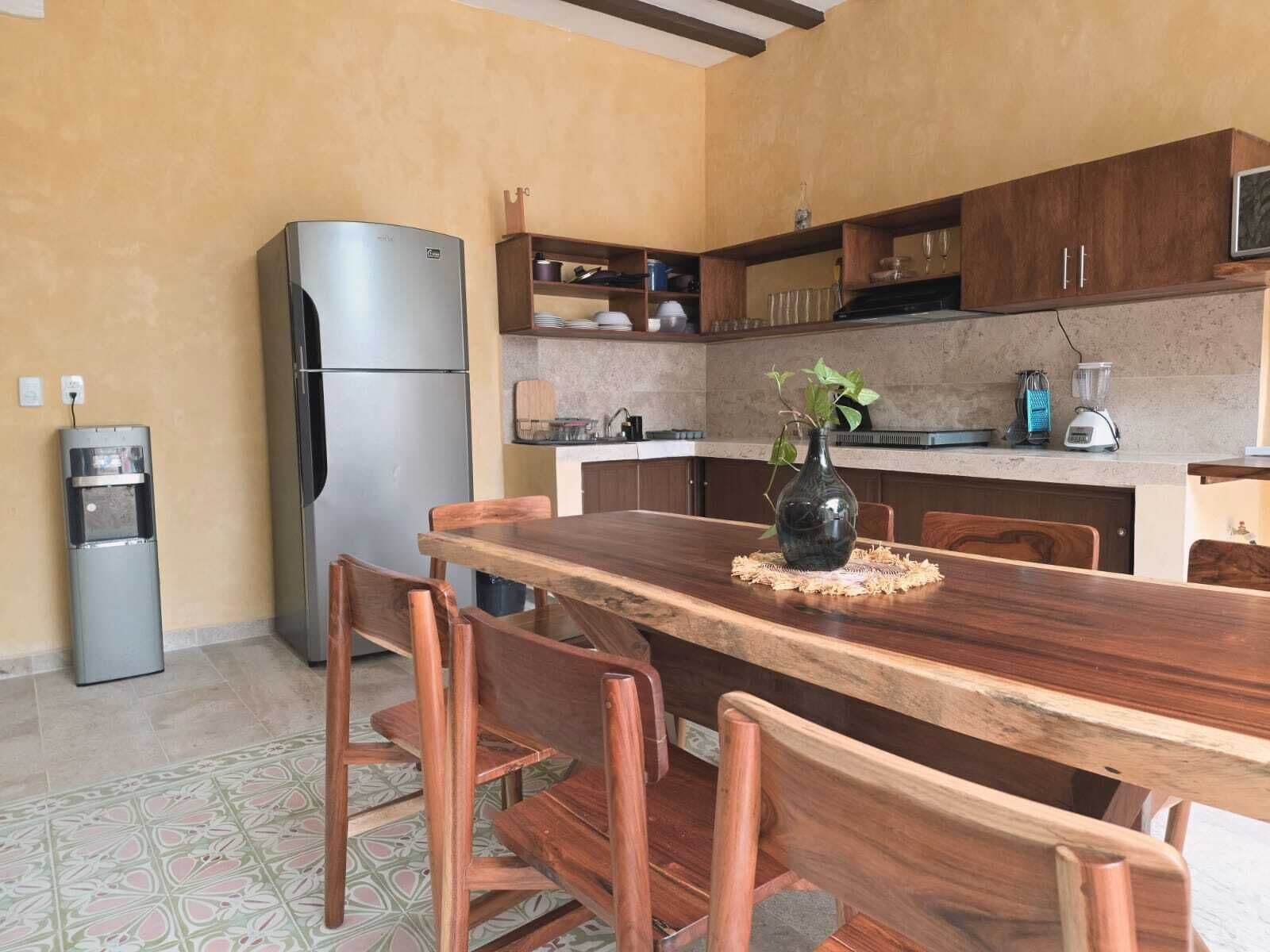 A compact kitchen with a stainless steel refrigerator, a water dispenser, wooden chairs, a dining table, and a backsplash with tiles.