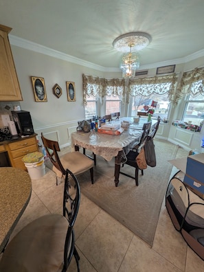 A dining area with a table covered by a lace tablecloth, surrounded by chairs. There are framed pictures on the wall, a chandelier, and a view of the outside through large windows. - Bunkbed In Quiet, Friendly Family-Hosted Home in South Jordan Near Everything (South Jordan)