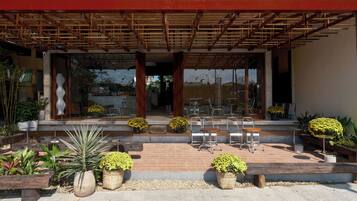 Un área de estar al aire libre con mesas y sillas, rodeada de plantas en macetas y una pérgola de madera.