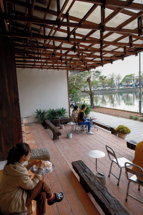 An outdoor seating area with wooden tables and chairs, a wooden pergola overhead, and a view of a lake with a boat dock. - Amea Homestay (Hue)