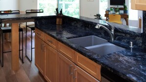 A modern kitchen with wooden cabinets, a black granite countertop, a double sink, and a dining area with a wooden table and chairs. - Aptos Hills Getaway - Welcome to Oak Landing (Aptos)
