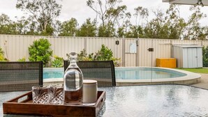 Un tavolo con ripiano in vetro su cui è posata una teglia con un barattolo di vetro, una tazza e due bicchieri, con sullo sfondo una piscina e un recinto alberato.