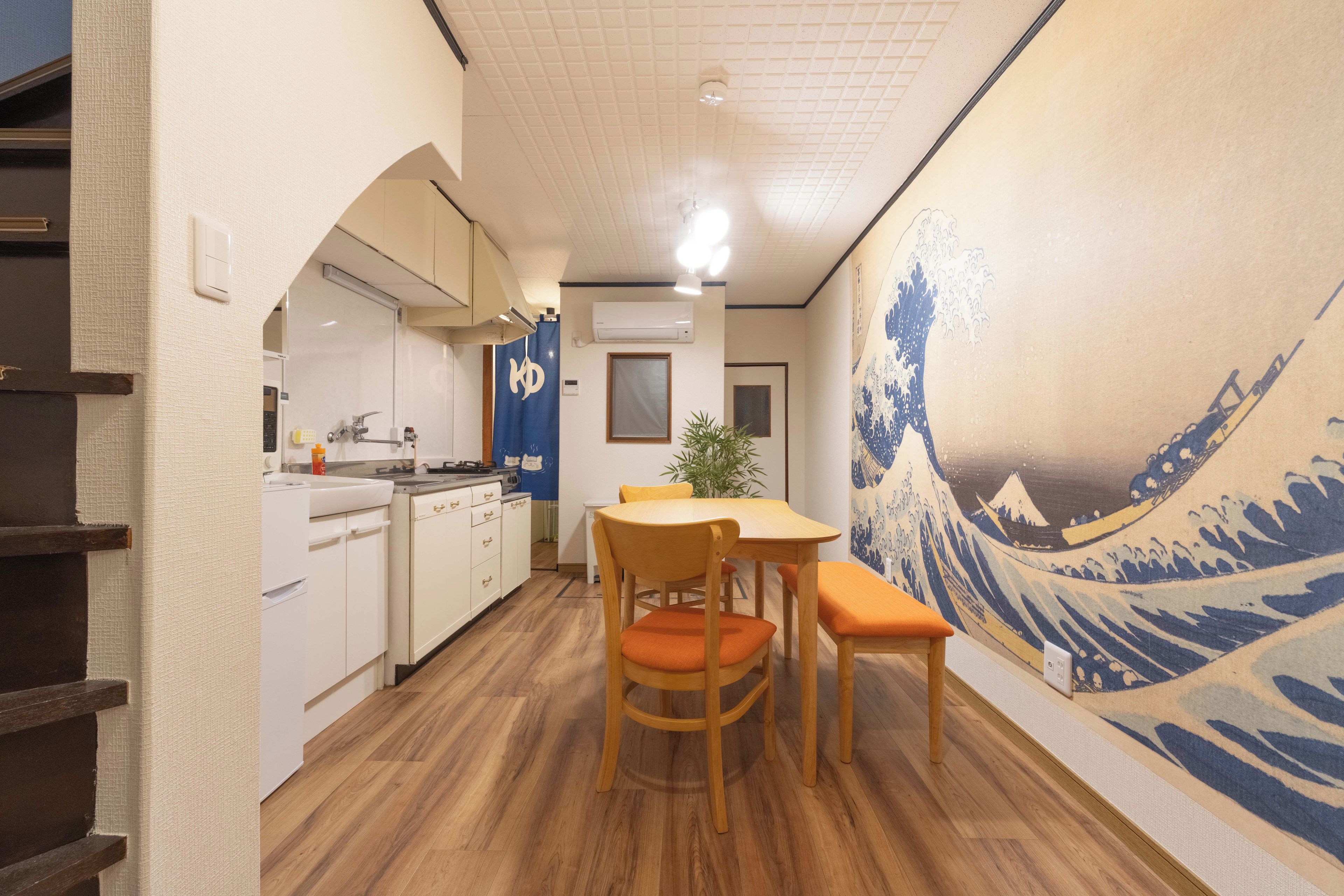 A compact kitchen with white cabinets, a sink, and a stove. There is a dining area with a table and chairs, and a wall with a wave painting.