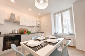 A modern kitchen with white cabinets, a stove, and a dining area set for two with a table, chairs, and a window with curtains. - Quarda, Apartment in Savona Old Town, Savona, Italy (Savona)