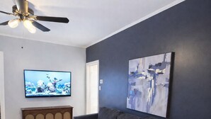 Ein Wohnzimmer mit einem dunkelgrauen Sofa, einem Holztisch, einem Deckenventilator, einem Flachbildfernseher und abstrakter Kunst an der Wand.