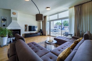 Ein modernes Wohnzimmer mit einer großen Sofagarnitur, einem Flachbildfernseher, einem Kamin und Blick ins Freie durch große Fenster.