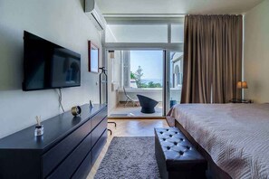Ein modernes Hotelzimmer mit Flachbildfernseher, dunklem Holzschrank, einem breiten Bett mit gepolstertem Kopfteil, einer kleinen Bank und Blick durch eine Glastür nach draußen.