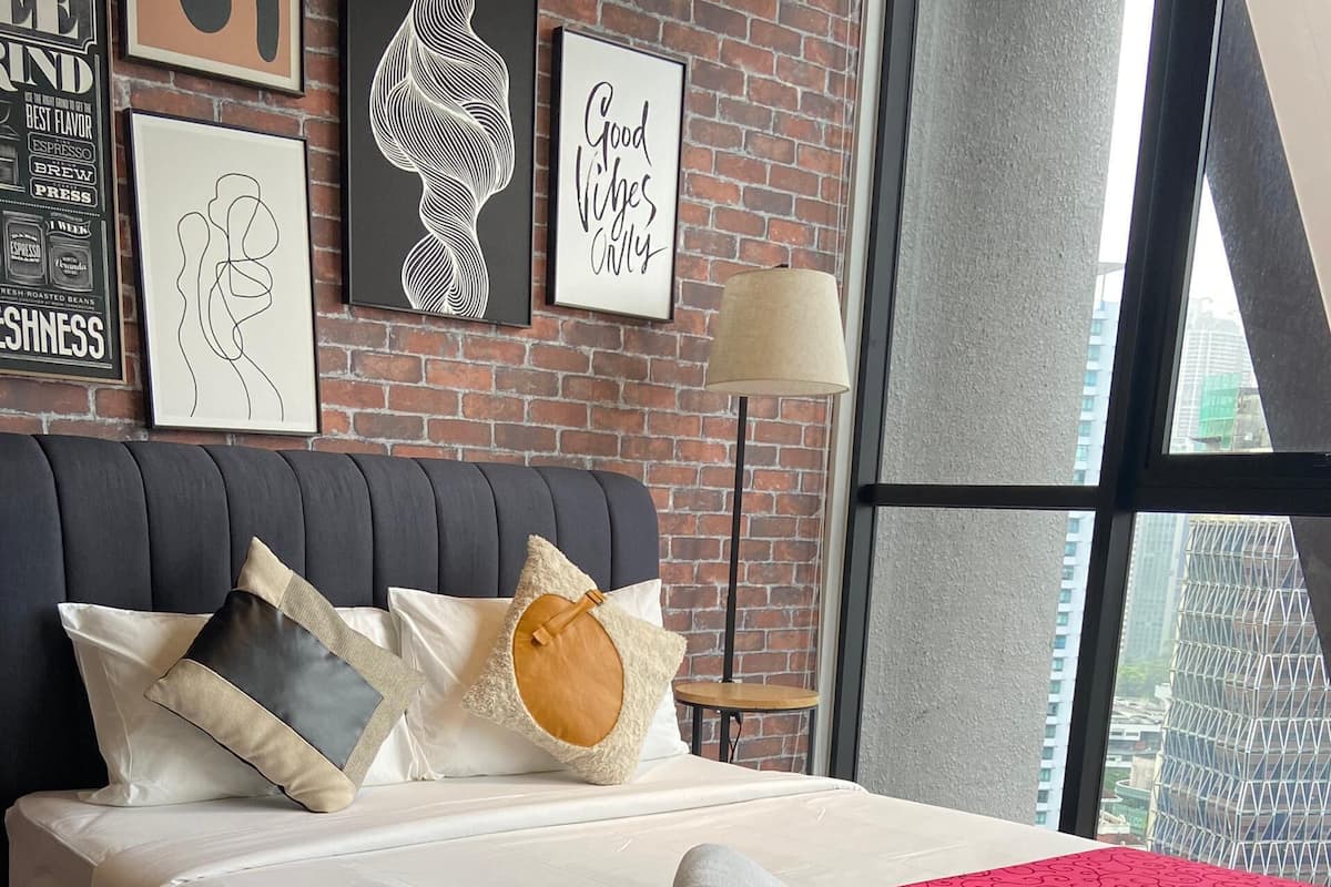 Quarto de hotel moderno com parede de tijolos, uma cama grande com várias almofadas, um cobertor vermelho e vista para o horizonte urbano através de janelas amplas.