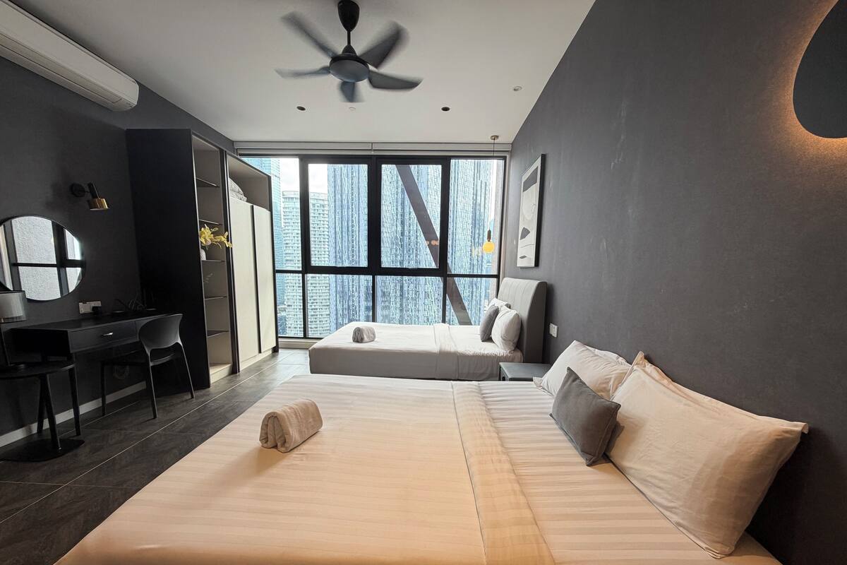 Quarto de hotel moderno com uma cama grande, uma escrivaninha e vista para edifícios altos através de grandes janelas.