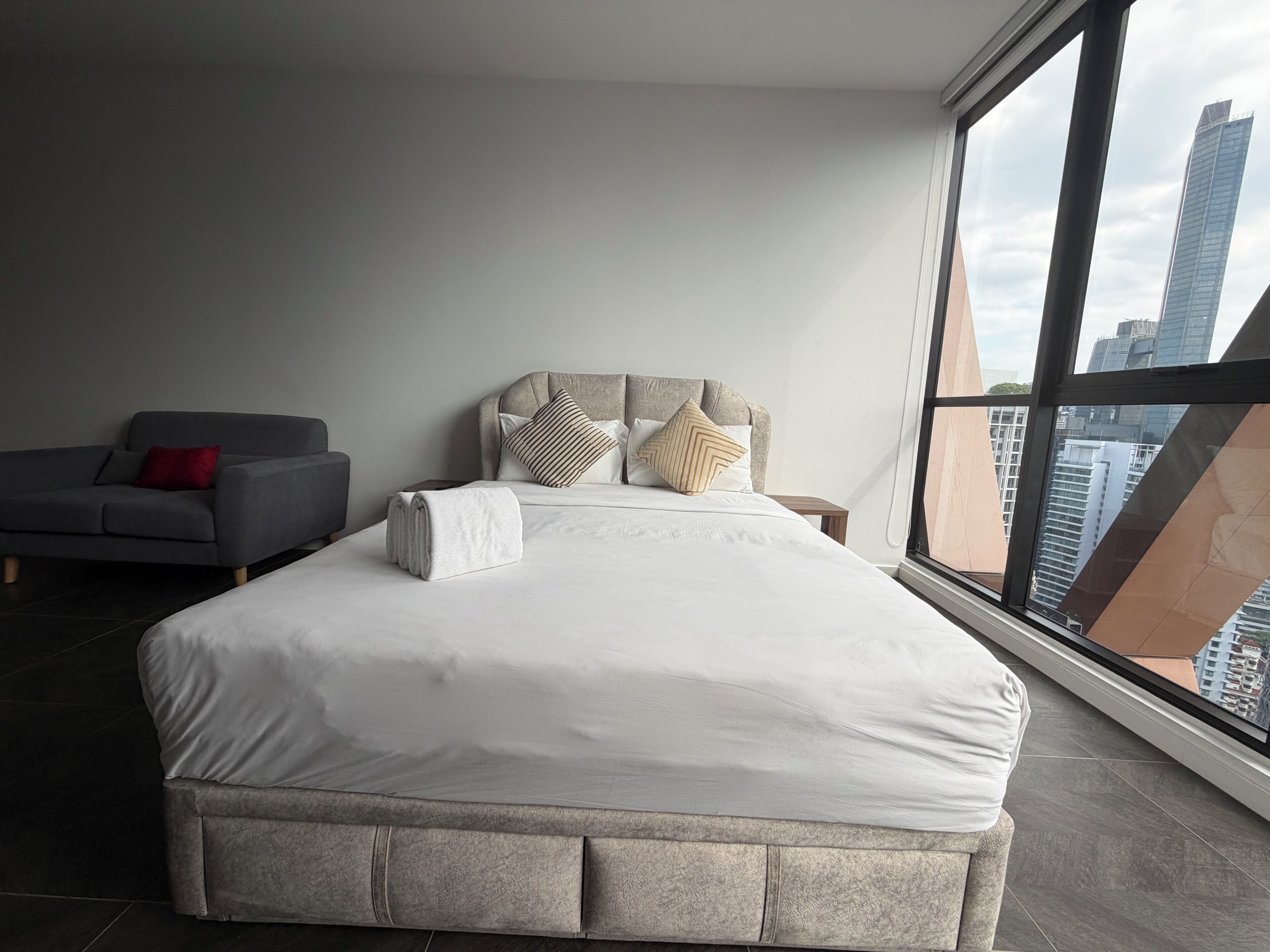 Ein modernes Hotelzimmer mit einem großen Bett, einem grauen Sofa und Blick auf die Stadt durch ein großes Fenster.
