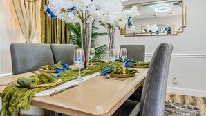 Mesa de jantar posta com guardanapos verdes, talheres e copos de vinho, rodeada por cadeiras estofadas cinzentas.