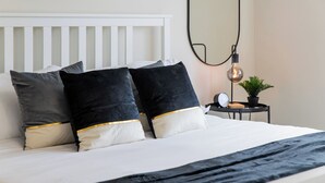 Una cama bien hecha con cabecera blanca, almohadas negras y doradas, y una manta negra. Hay una mesita de noche con un reloj, una lámpara y una planta en maceta.