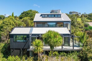Une maison moderne à deux étages, dotée d’un toit plat, de grandes fenêtres et d’une terrasse entourée d’une végétation luxuriante.