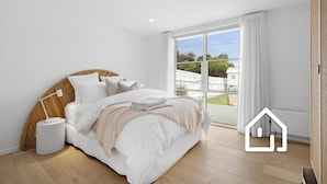 Een slaapkamer met een groot bed, een bedlamp, een rond rieten bijzettafeltje en uitzicht buiten door een groot raam.