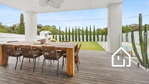 Een terras buiten met een houten tafel en stoelen, een plafondventilator en uitzicht op een zwembad en cactussen.