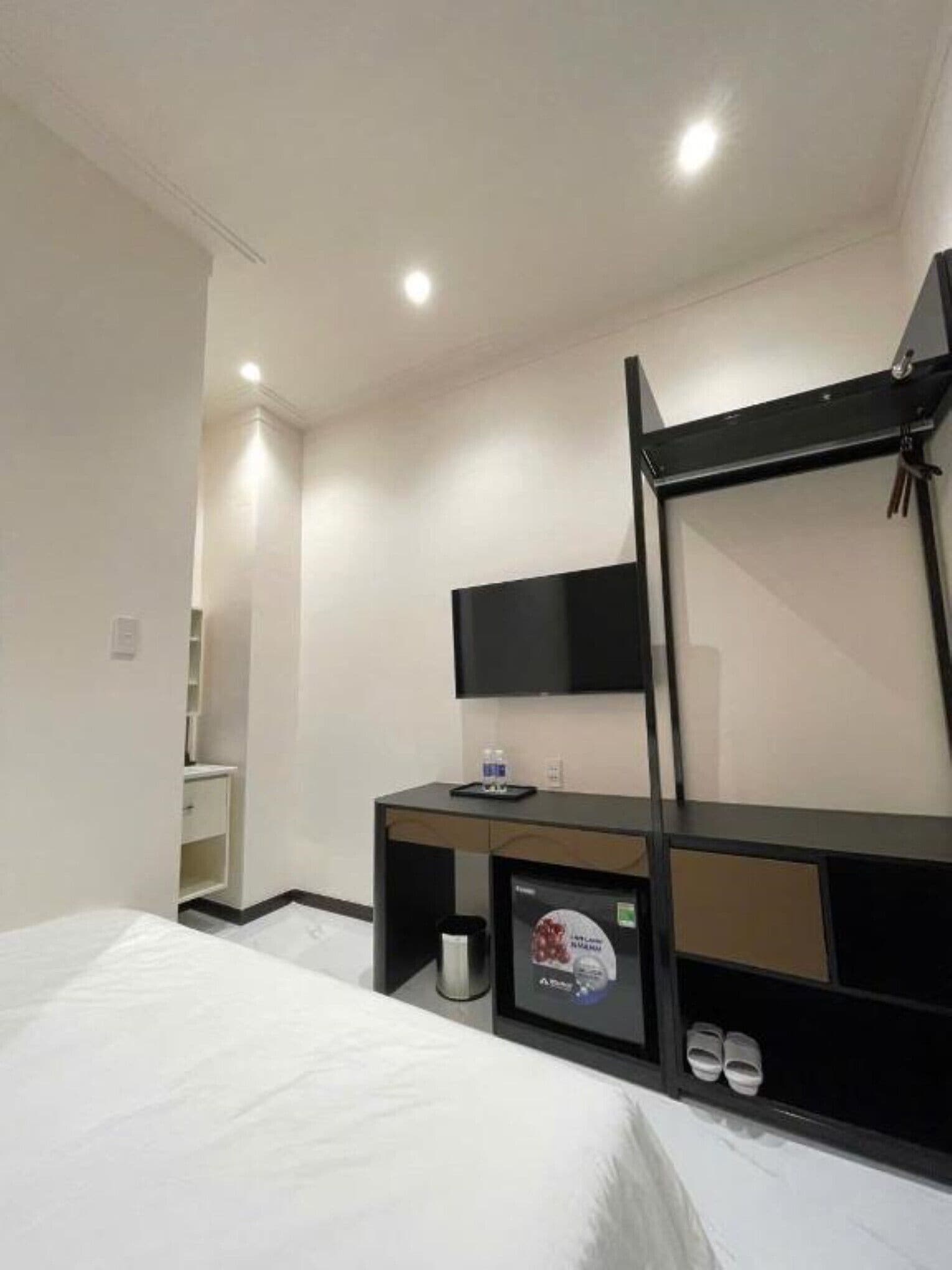 A hotel room with a bed, a desk with a flat-screen TV, a small shelf with a water bottle, and a trash bin.