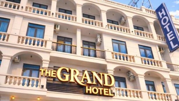 Un hotel de varios pisos con un llamativo letrero que dice 'The Grand Hotel', ventanas amplias y un balcón en los pisos superiores.