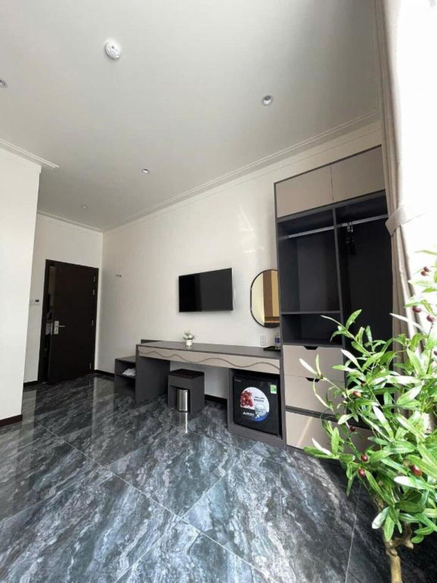 Habitación de hotel moderna con suelo de mármol, televisor de pantalla plana en la pared, un escritorio pequeño con silla y una planta en maceta.