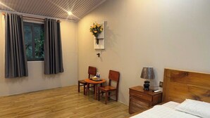 Habitación de hotel con piso de madera, cama con sábanas blancas, mesita de noche con lámpara, una pequeña mesa con sillas y ventana con cortinas.