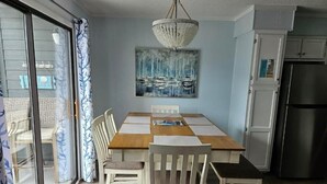 Un comedor con una mesa de madera y cuatro sillas, una lámpara de araña, un cuadro de barcos en la pared y un puerta corrediza de vidrio que da a un espacio exterior.