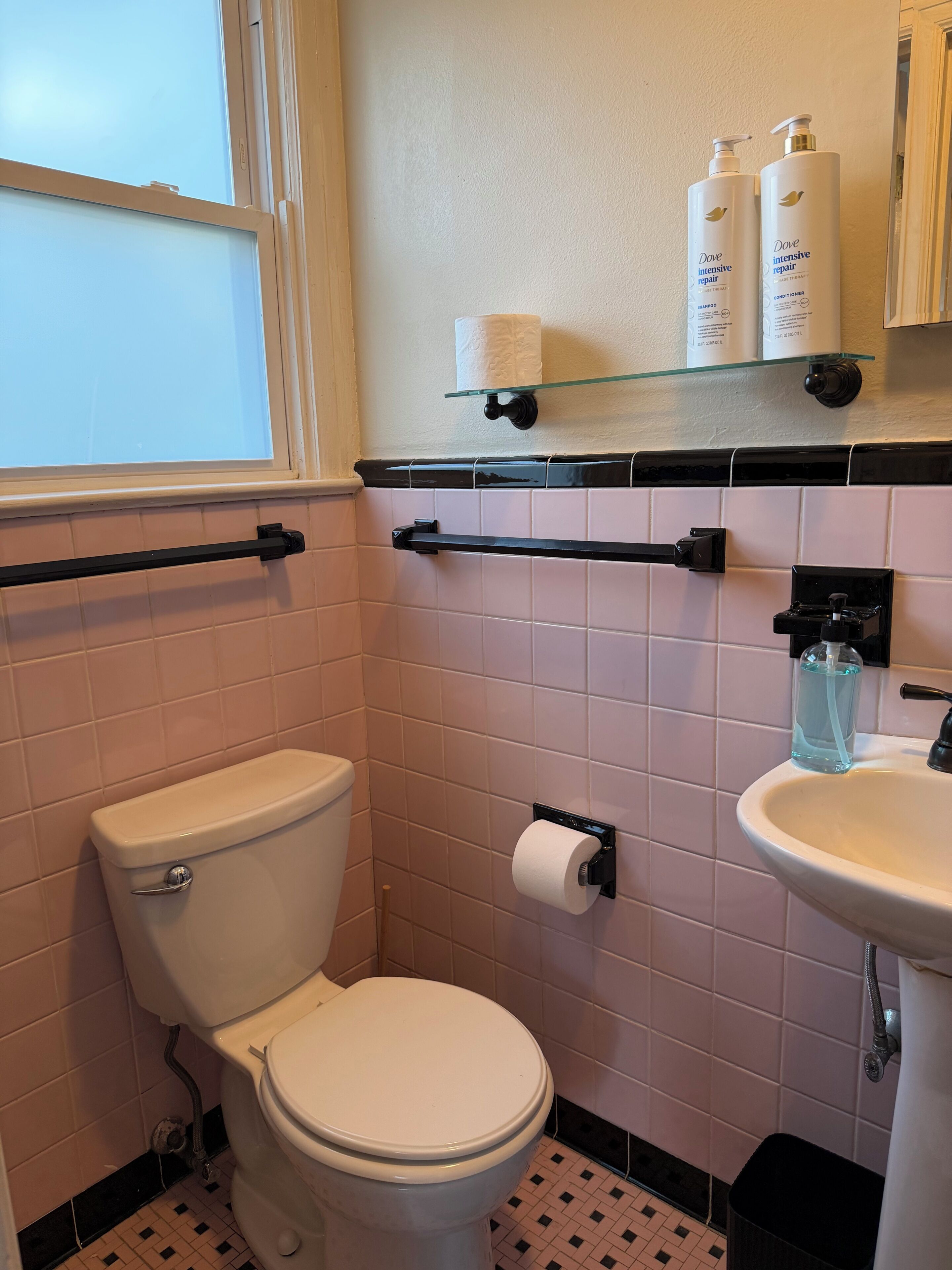 Ein Badezimmer mit Toilette, Waschbecken, Ablagefläche aus Glas mit Pflegeprodukten und einem Fenster.