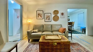 A living room with a sofa, a coffee table, and a floor lamp. There are framed pictures on the wall and a dining area in the background. - Location! Charming neighborhood Newly renovated Modern furnishings  Fab Staycay (Fort Worth)