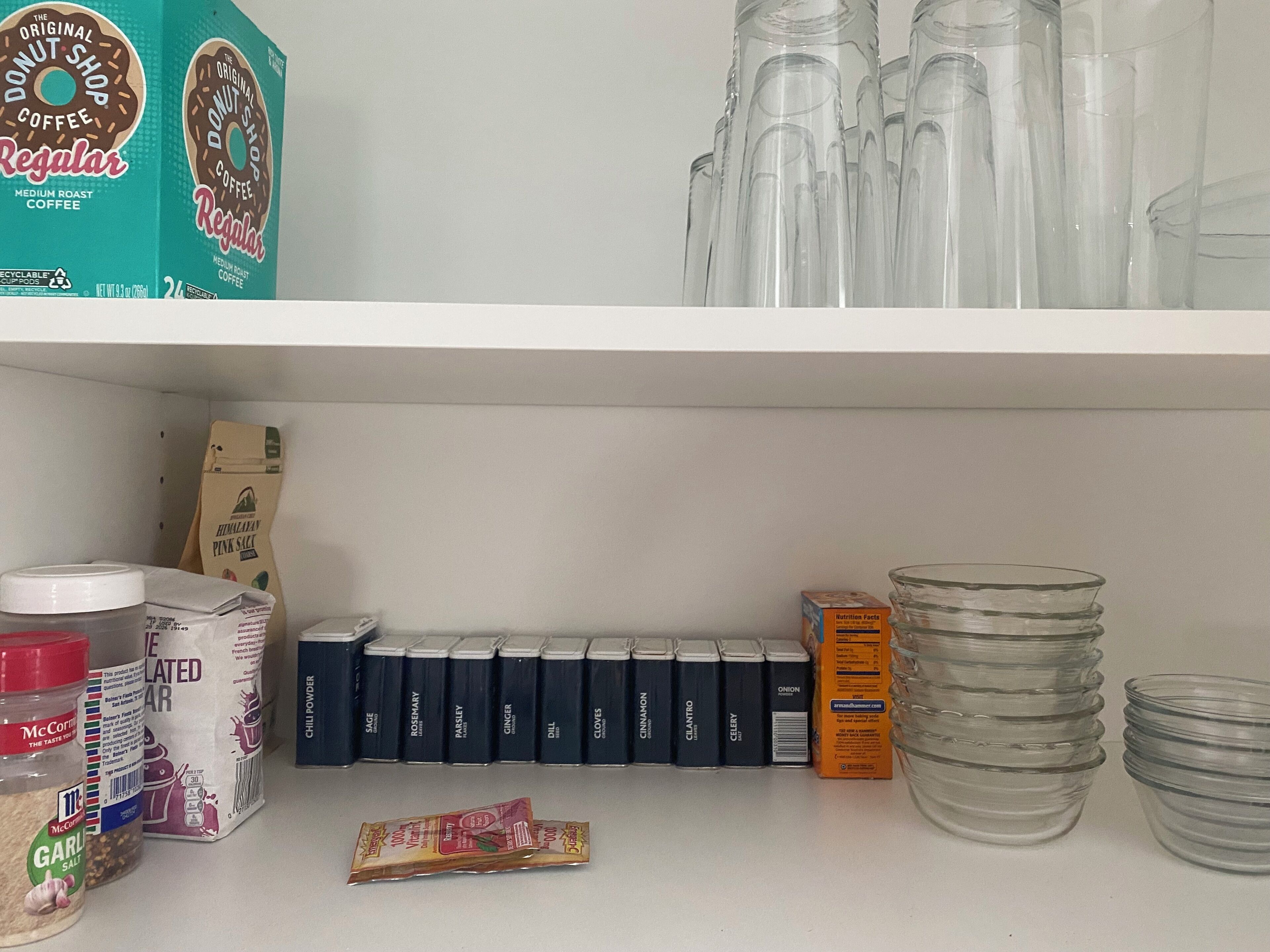 A kitchen shelf with various items including a box of The Donut Shop Coffee, a bag of Himalayan Pink Salt, McCormick spices, and a stack of clear glass cups.